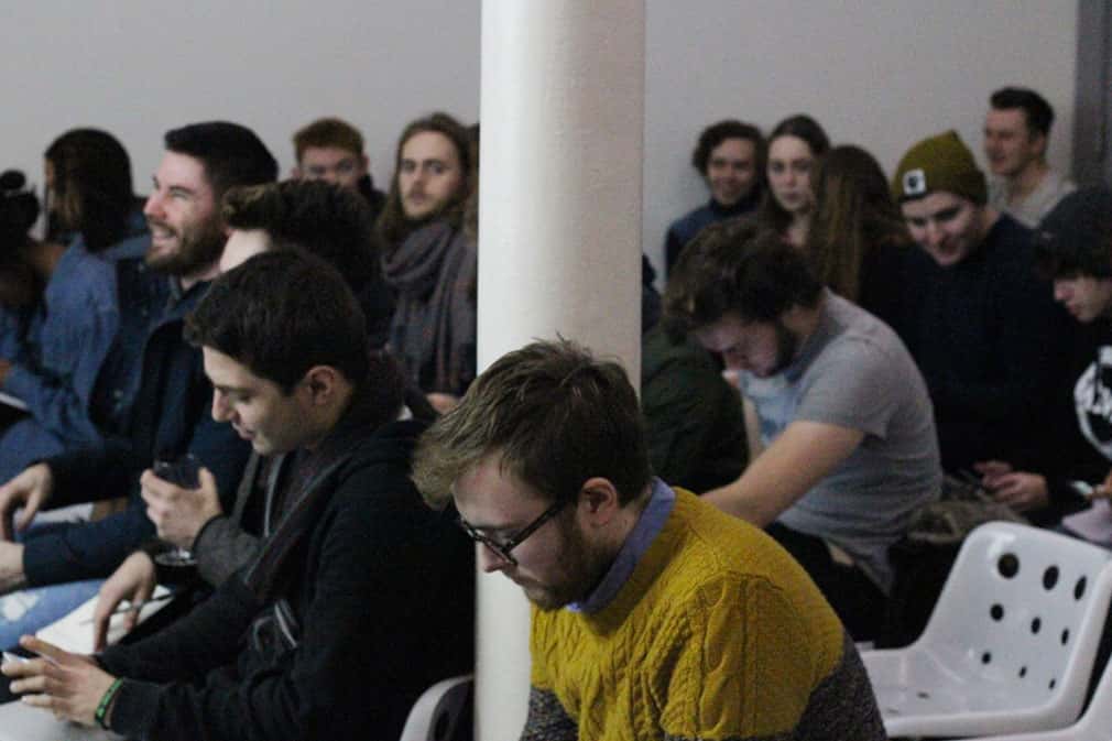 photo of students sitting in white plastic chairs with several writing notes and a white column in the centre of the room