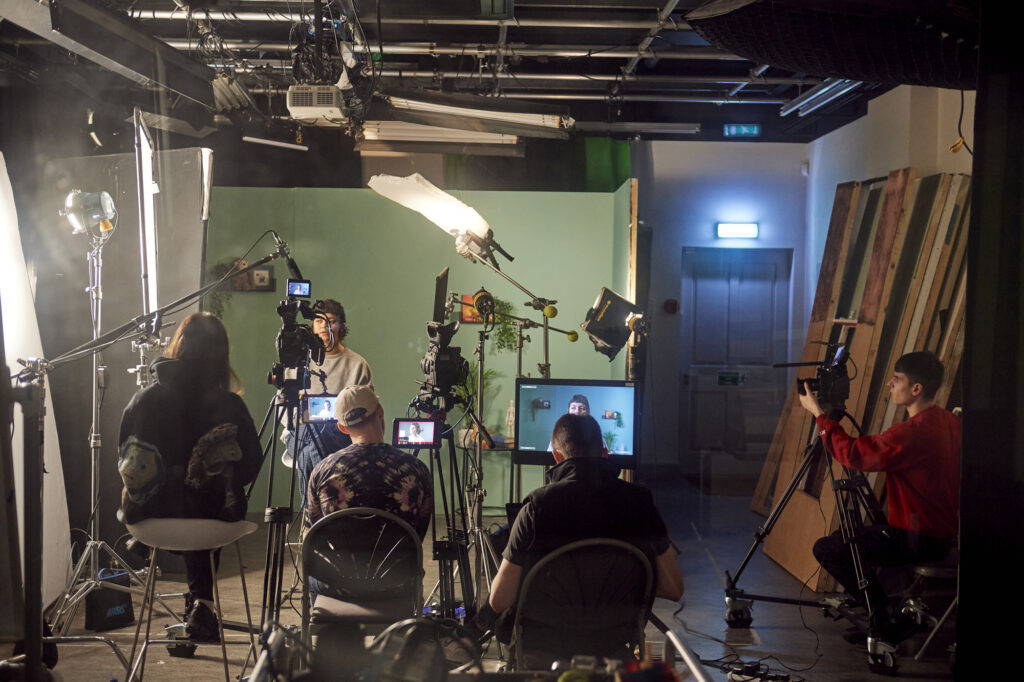 A behind-the-scenes view of a film or video shoot shows a crew recording a person seated under studio lights—an experience similar to what students gain while pursuing an MA Moving Image and Sound degree, with cameras, crew, and equipment on set.