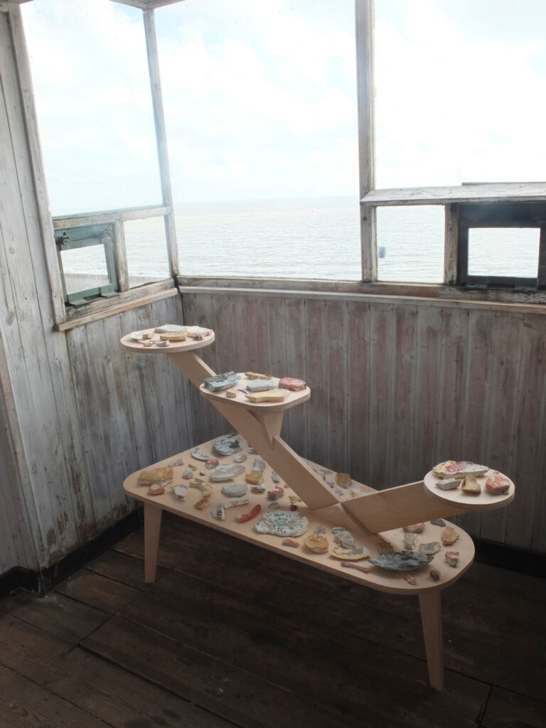 A wooden tiered display table holding various small ceramic pieces stands in a weathered, sunlit room with peeling white walls and windows overlooking a body of water.