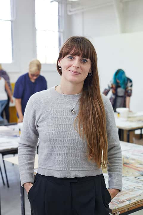 portrait photo of lecturer Sarah Longworth-West standing with hands in pockets and smiling at camera with long brown hair and a long sleeved patterned knit jumper