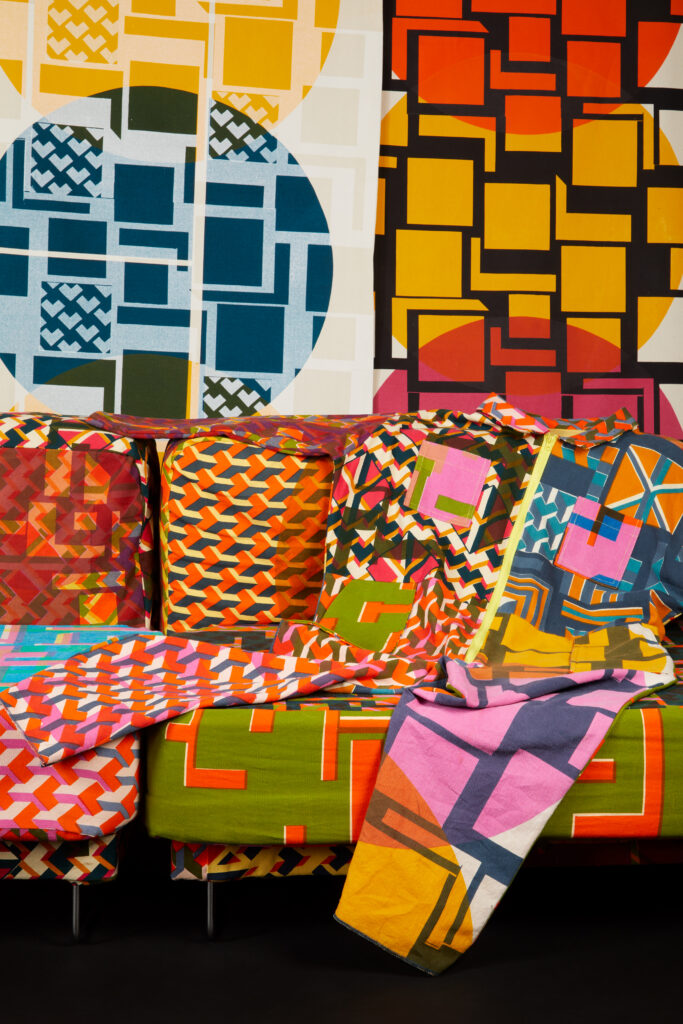 A colorful couch and blankets with bold geometric patterns, reflecting the creativity of a textile design degree, sit in front of a wall decorated with vibrant square and circular designs in red, orange, yellow, blue, green, and pink.