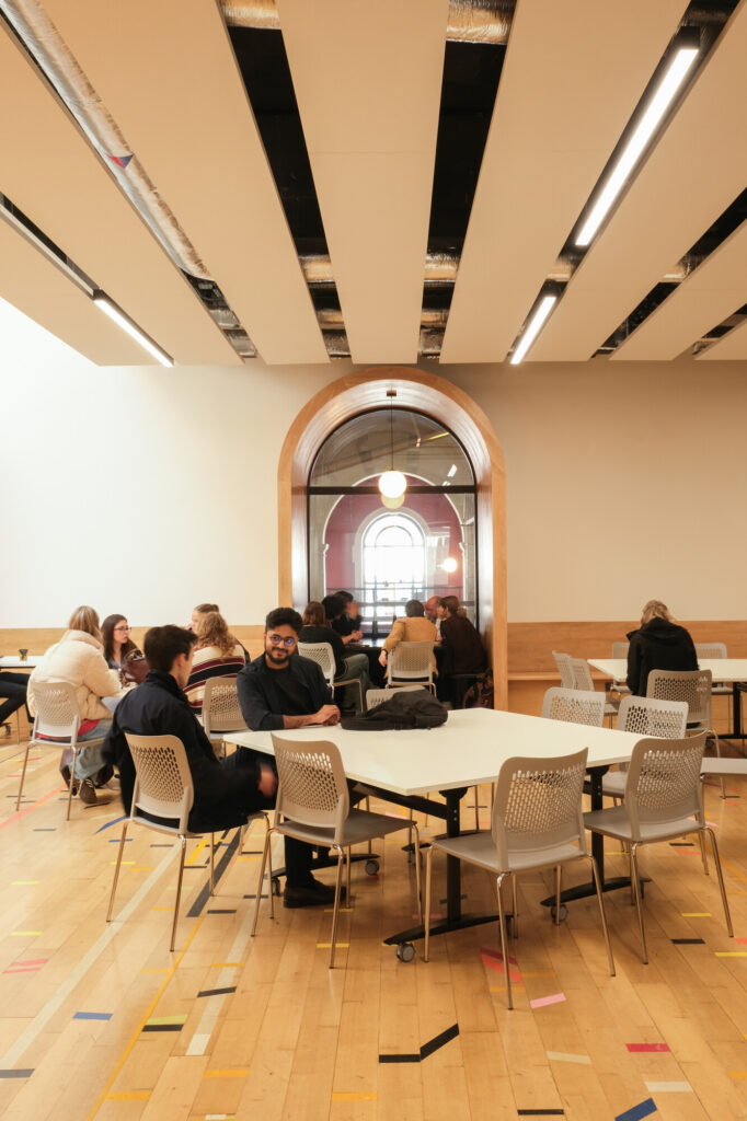 A group of people sit and talk around tables in a spacious, modern room with wooden floors, white walls, and a large arched window in the background letting in light.