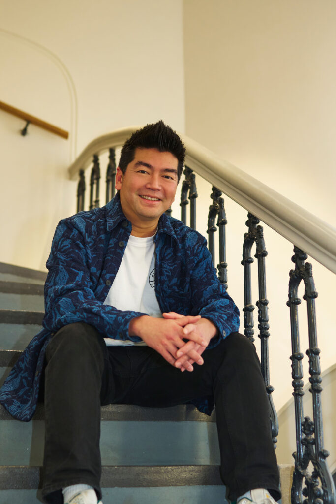A man with short dark hair, wearing a blue patterned shirt over a white t-shirt, black pants, and white shoes, sits smiling on an indoor staircase with black railings.