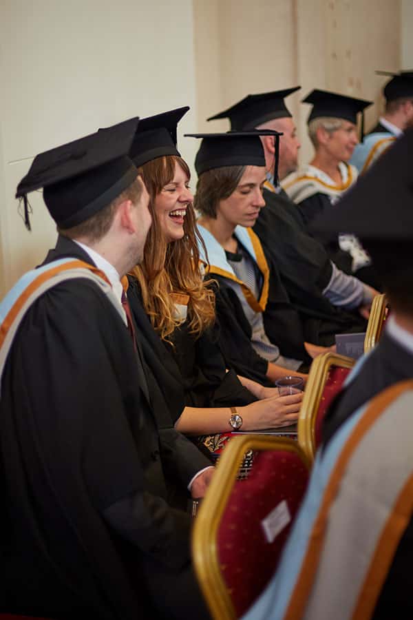Students sat in a Norwich University of the Arts graduation in a row, smiling and laughing wearing their caps and gowns