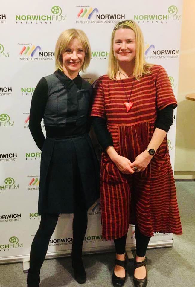 Two women stand and smile in front of a backdrop with "Norwich Film Festival" and "Norwich Business Improvement District" logos. One wears a black dress; the other wears a red striped dress with a heart necklace.