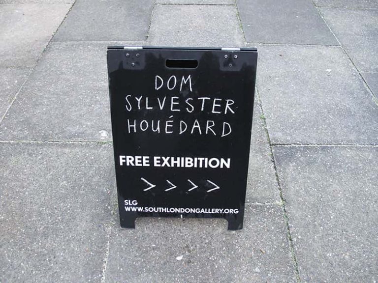 concrete floor with black standing chalkboard in the middle saying Dom Sylvester Houédard, free exhibition. SLG www.southlondongallery.org