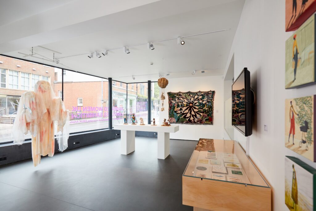 A bright contemporary art gallery with large windows, featuring sculptures on white pedestals, colorful wall hangings, paintings, a display case with documents, and a large textile figure near the window.