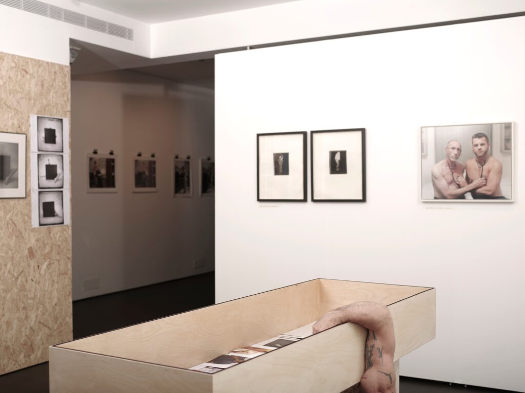 A contemporary art gallery with framed photographs on white walls. In the foreground, an arm hangs out of a wooden box, partially visible, suggesting a person inside. The space appears minimal and modern.