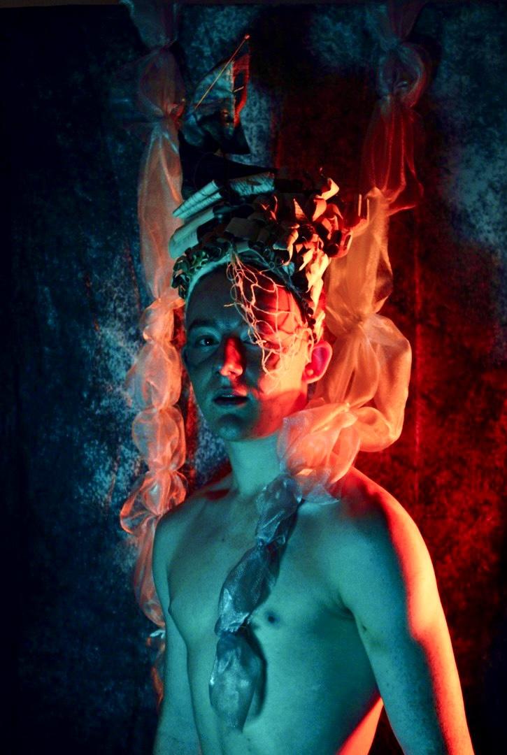Photograph of a person; they have no top and are wearing a strange hat. They are dramatically lit with red and blue lights from below.