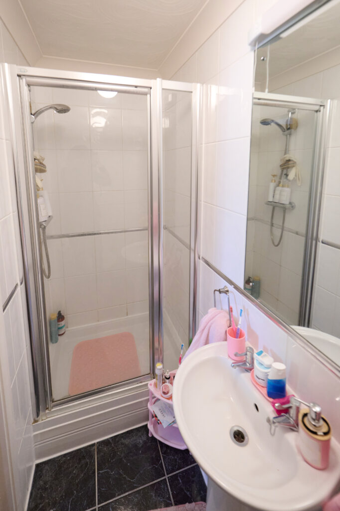 A small bathroom with a glass-enclosed shower, white tiled walls, a pink shower mat, and shelves with toiletries. Next to the shower is a white sink with soap, toothbrushes, and toothpaste on the counter.