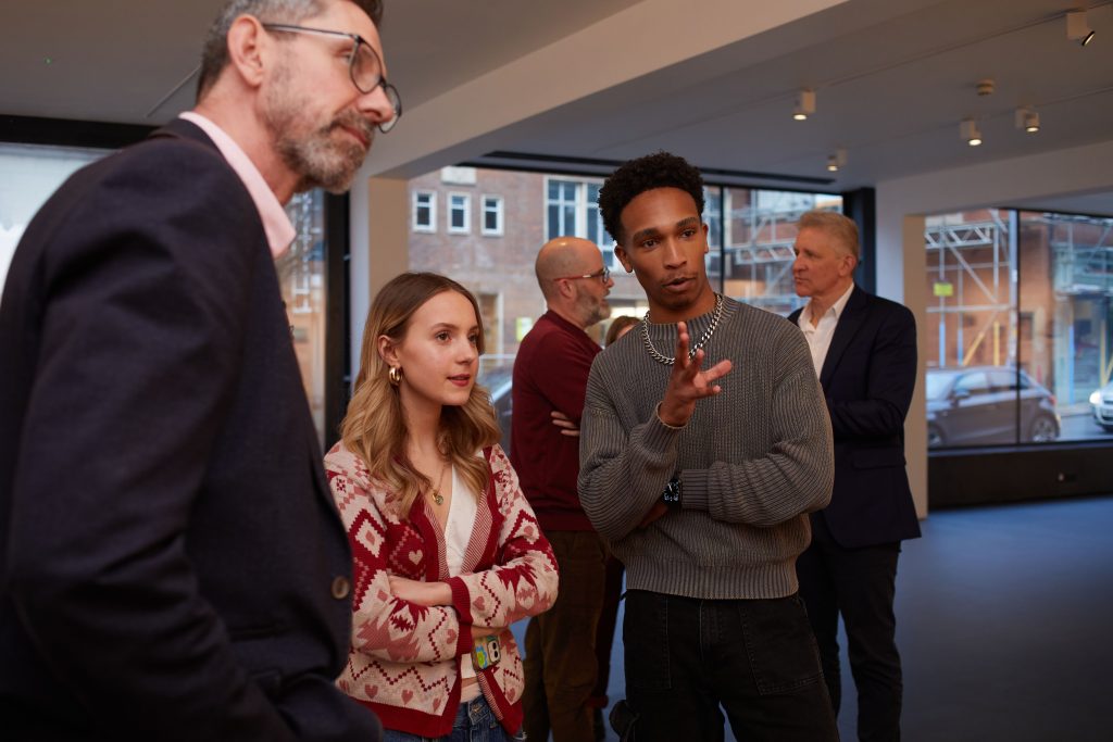 Three people gathered together discussing something in a gallery. One person is gesturing with their left hand.