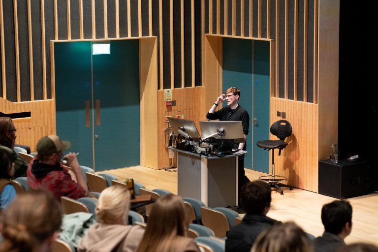 A man in a black shirt stands at a podium in a modern auditorium, addressing an attentive audience seated in rows.