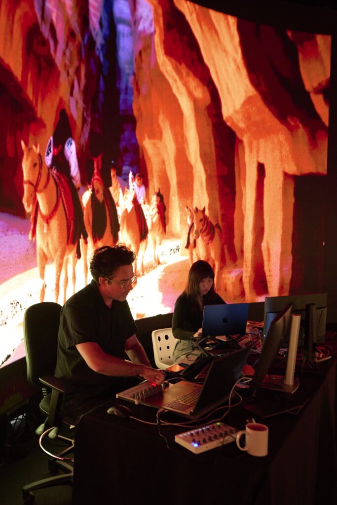 Two people operate computers and audio equipment in a dark room, with a large vibrant screen behind them showing an animated scene of horses and people traveling through a red and orange canyon.