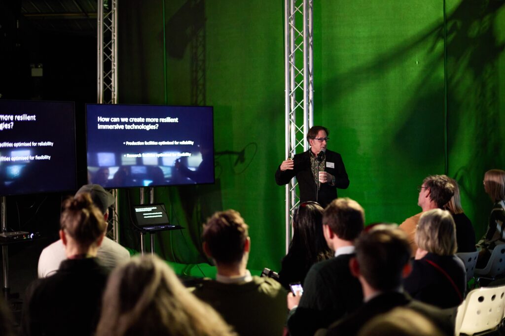 A speaker stands in front of a seated audience, gesturing towards a large screen displaying a presentation about immersive technologies, in a room with green lighting and metal truss structures.