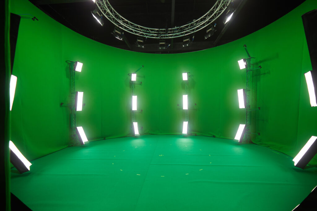 A large green screen studio setup with bright vertical lights arranged in a semi-circle around the space and a circular metal structure overhead.