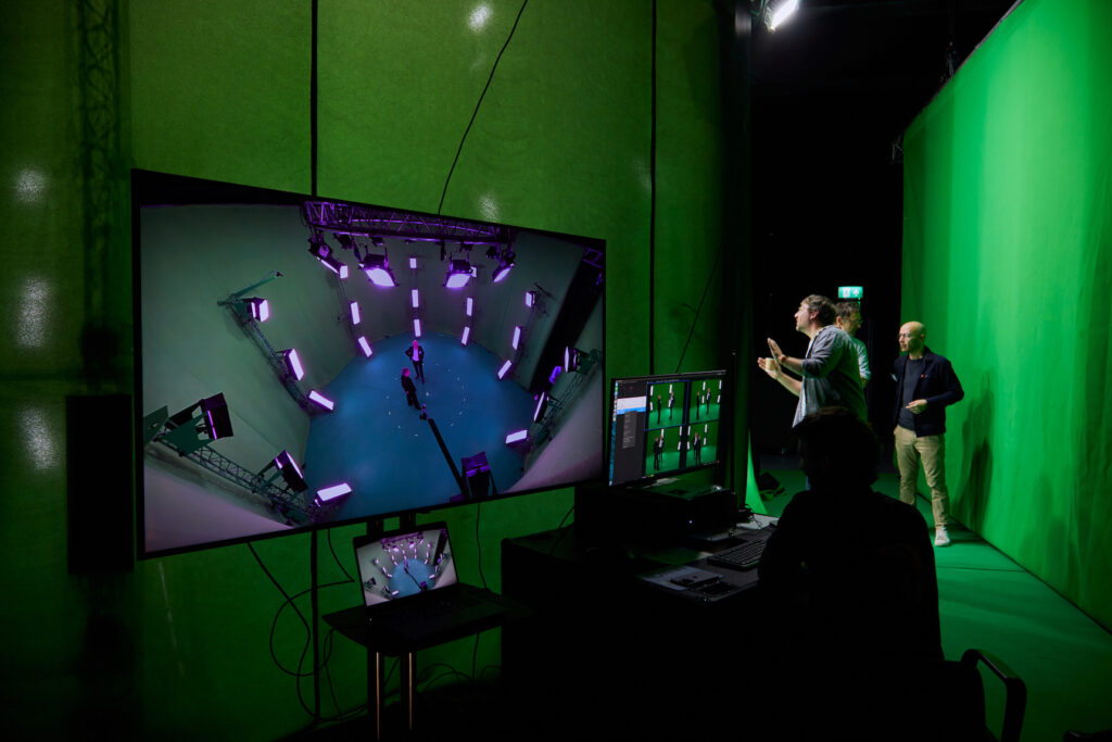 Three people work in a studio with green screens and multiple monitors. One monitor displays a bird’s-eye view of a person in a high-tech setup surrounded by lights, while another person operates equipment at a desk.