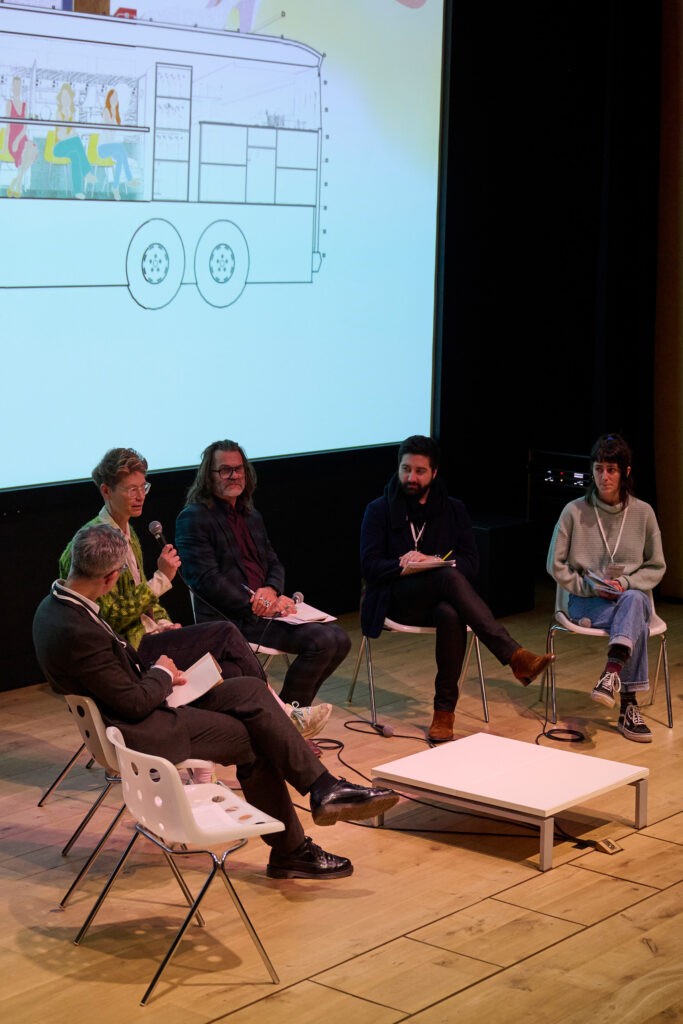Five people are seated in a semi-circle on stage having a panel discussion, with one person speaking into a microphone. A screen behind them shows an illustration of a bus, suggesting the event may explore themes in architectural humanities.
