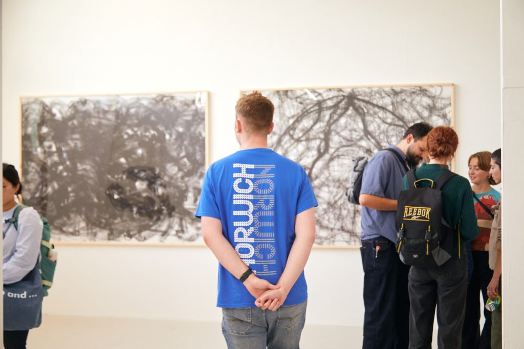 Ambassador standing in Grad Fest during degree shows, his back is facing the camera, blue Norwich Ambassador top. Visitors on either side of him.