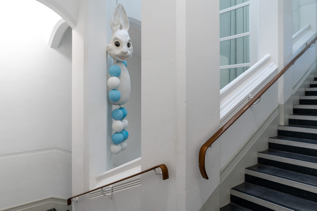 A photograph from PG Fest. In the stairwell of St Georges Building, a giant bunny sculpture is suspended from a gap in the wall. The bunny has a giant head with it's ears pointing upwards, tapering to meet at the tips. The torso and limbs are made of blue and white circular sculpted balls.
