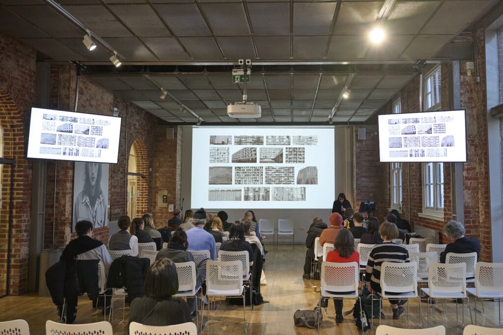 People sit facing a presenter in a brick-walled room with two large display screens and a projection of images on the front wall. The audience watches attentively, and the atmosphere is that of a formal lecture or presentation.