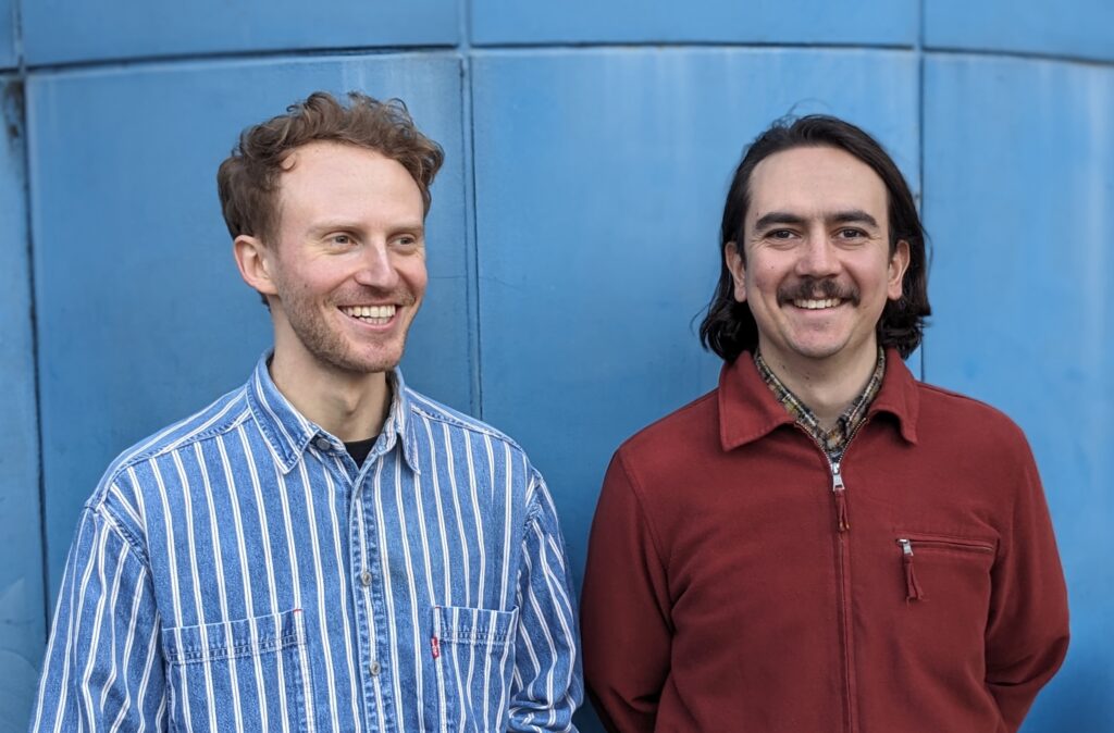 Two people are standing in front of a blue wall. The person on the left is wearing a blue and white striped shirt, while the person on the right is wearing a maroon jacket. Both are smiling and looking slightly to the side.