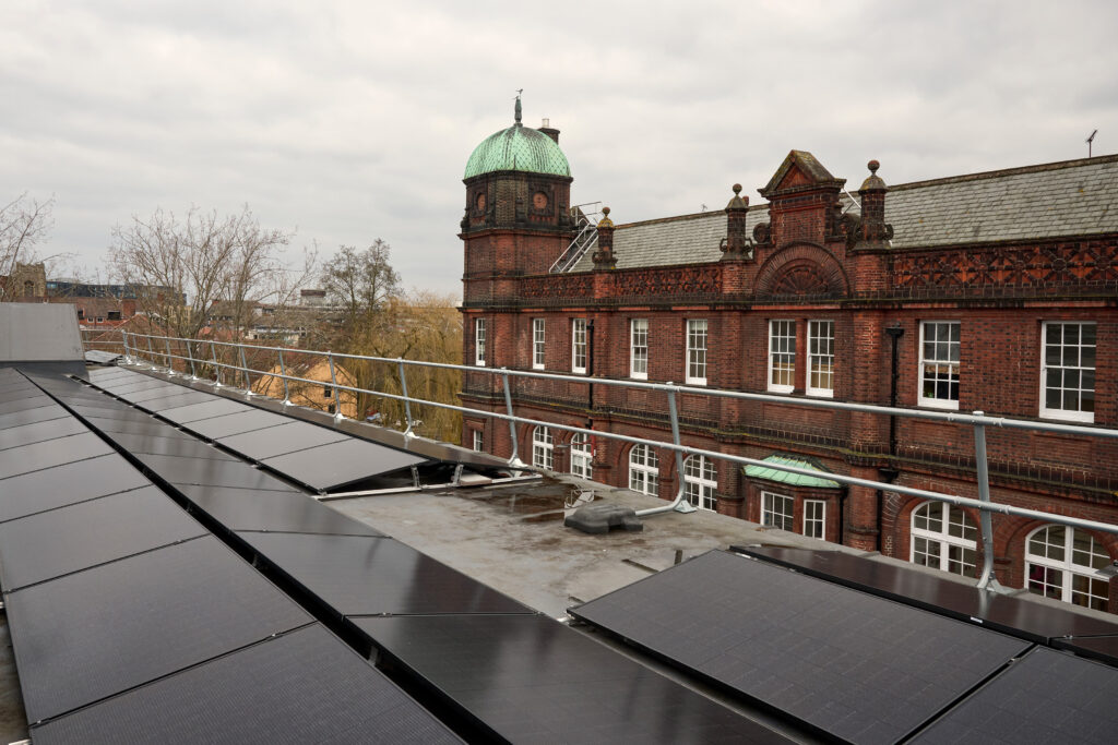 Rooftop solar panels with a historic red-brick building in the background. The building features a dome and ornate architectural details. Overcast sky and bare trees suggest a cooler season. Rails border the rooftop area.