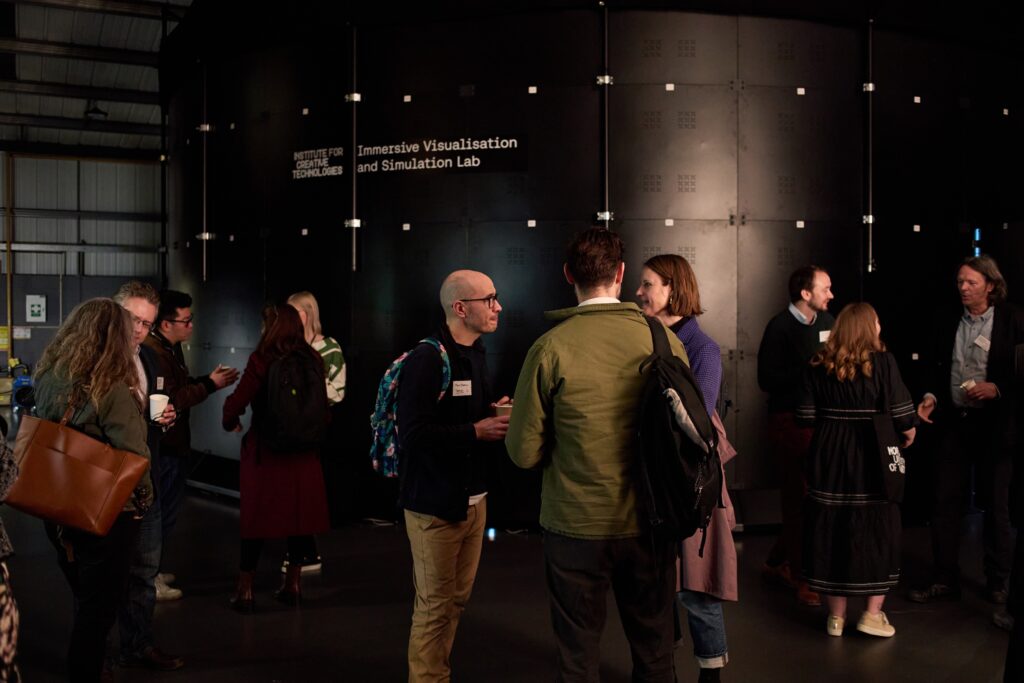 A group of people engaged in conversation inside a dimly lit room with a large black immersive visualization and simulation lab structure in the background. Some are holding drinks, and they appear to be at an informal event or gathering.