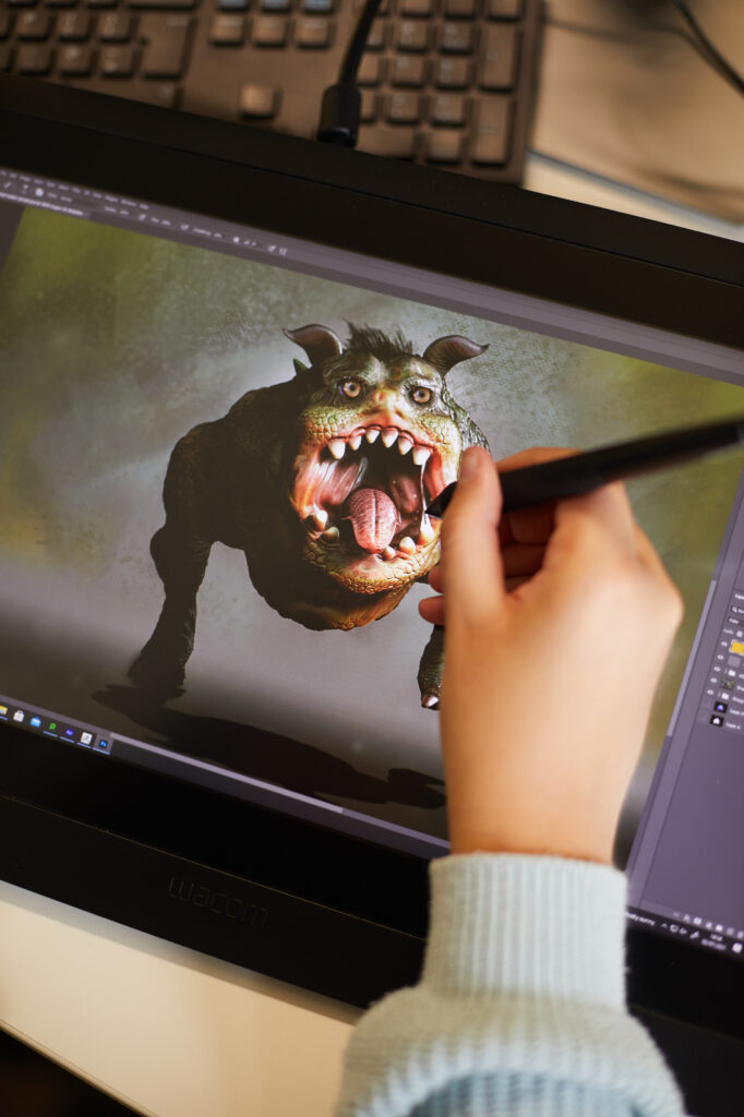 A person uses a stylus on a graphics tablet to create a digital image of a green, monstrous creature with large teeth and horns. The tablet displays detailed artwork, and a computer keyboard is visible in the background.