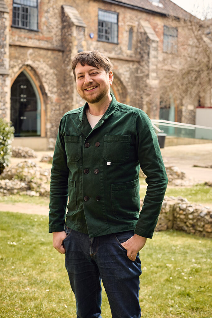 A man with short brown hair and a beard, wearing a green jacket and jeans, stands smiling outdoors on grass with historic stone buildings in the background.