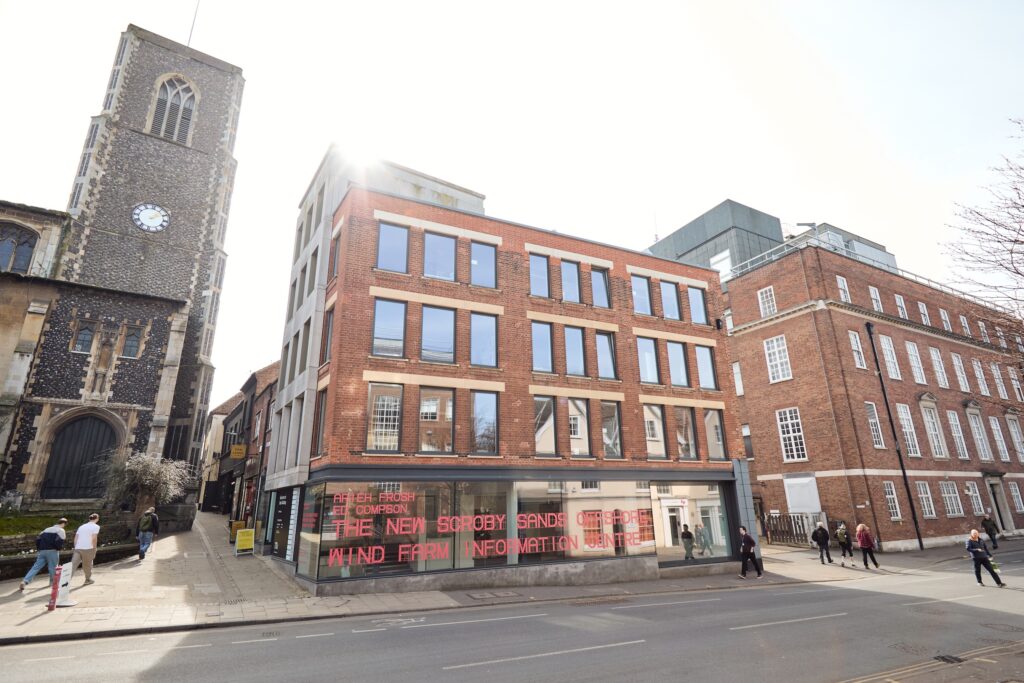A modern red-brick building with large windows sits on a city street, next to an old stone church with a clock tower. Pedestrians walk along the sidewalk under a bright sky.