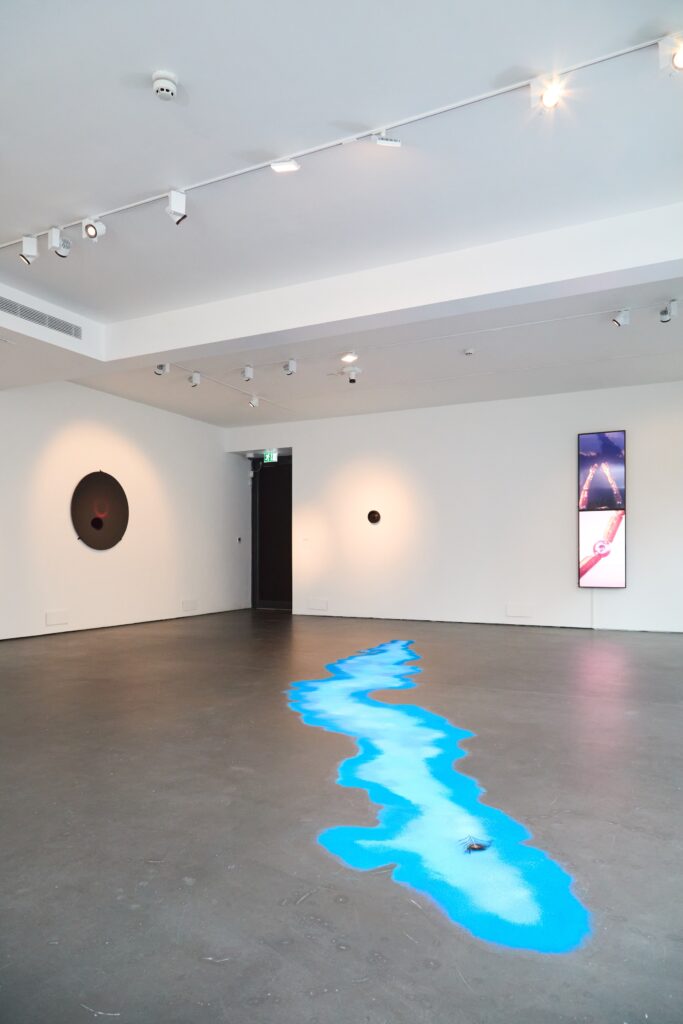 A modern art gallery with white walls features a blue, winding river-like installation on the gray floor. Two circular artworks hang on the walls, and a vertical digital screen displays abstract images. Ceiling lights illuminate the scene.