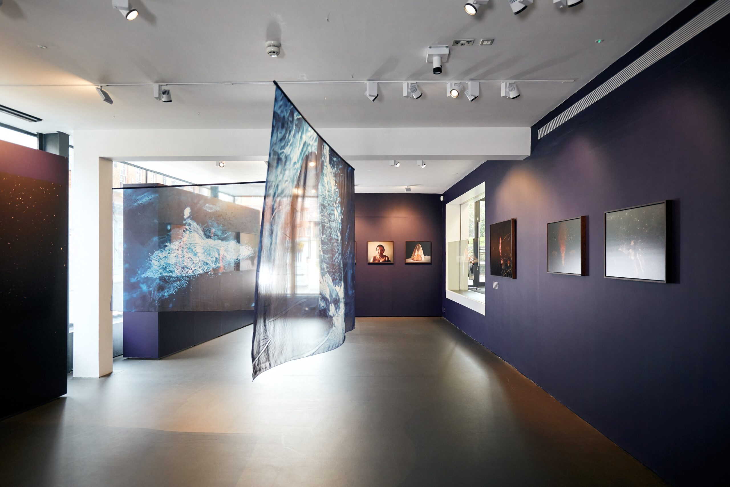 A contemporary art gallery with dark walls displays framed photographs and a large, semi-transparent fabric panel printed with a splash pattern, suspended in the center of the room. Natural light enters through windows on the right.