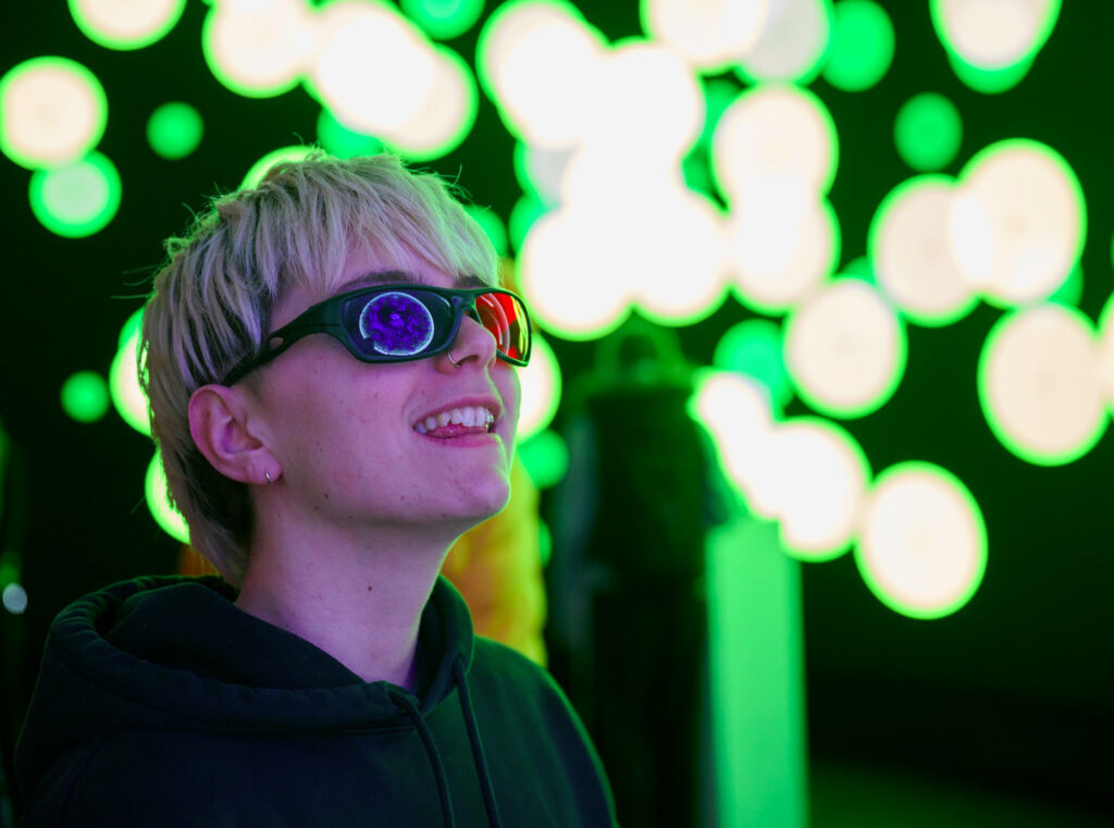 A person with short blond hair and sunglasses smiles while looking at bright, blurry green and white circular lights in the background.