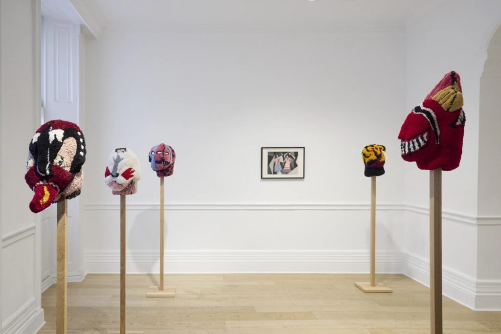 Five colorful, abstract textile masks are displayed on wooden stands in a white-walled gallery. A framed artwork hangs on the back wall above pale wooden flooring. The room is brightly lit.