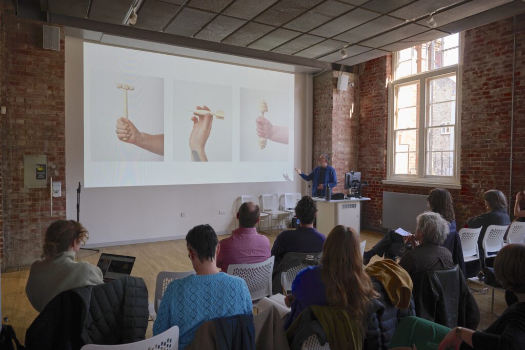 A presenter stands at the front of a brick-walled room, gesturing towards a screen displaying three images of hands holding objects, while an audience sits and listens attentively.