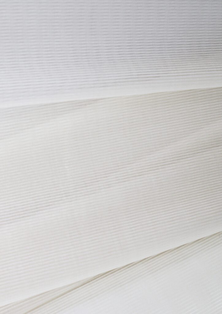 Close-up of several layers of thin, white and off-white fabric or paper with fine horizontal lines and subtle texture, creating a soft, minimal, and slightly striped appearance.