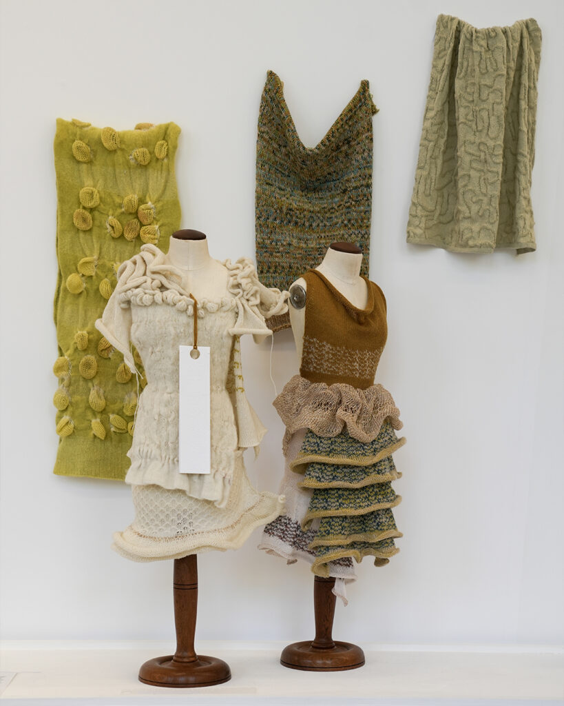 Two small mannequins display intricate knit dresses in cream and earthy tones. Behind them, three textured textile samples in yellow, green and beige shades hang on a white wall.