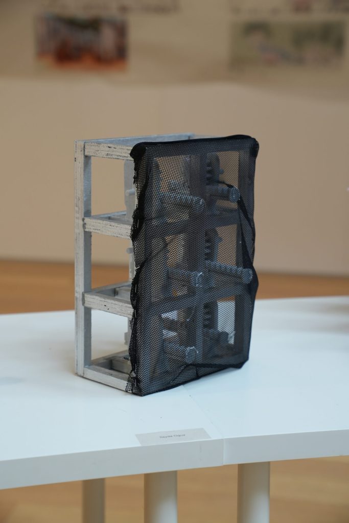 A rectangular metal sculpture covered partially with black mesh sits on a white display table.