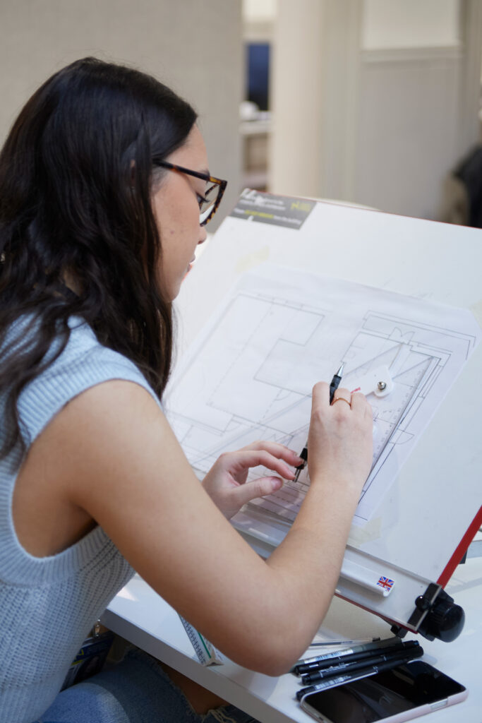 A person with long dark hair and glasses is clearing space on a drafting table to draw architectural plans using pens and rulers. Various drawing tools and a phone are placed nearby.