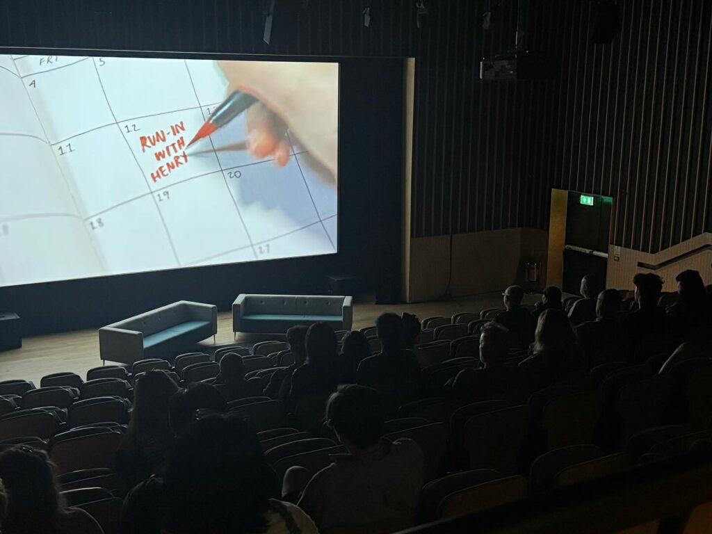 Audience seated in a dark movie theater watching a large screen that shows a hand writing “RUN IN WITH HENRY” on a calendar. The theater features modern seating and a couple of couches near the screen.