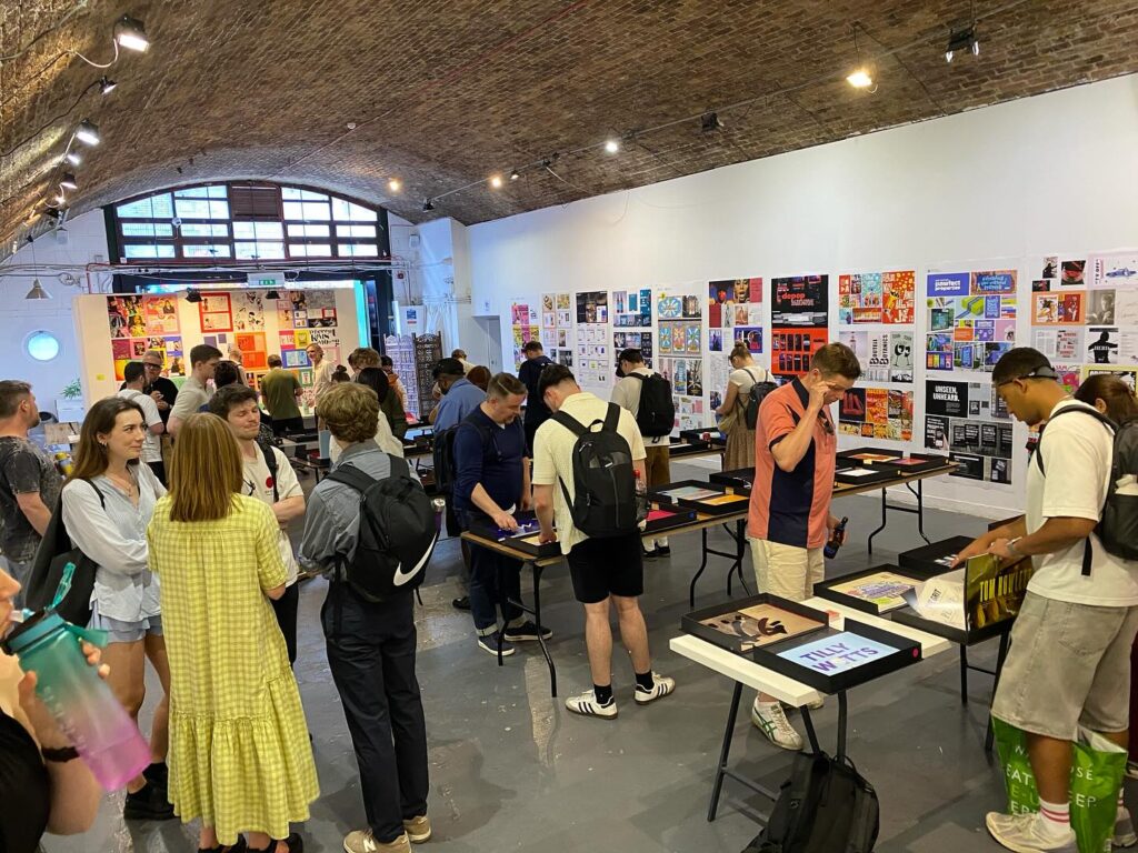 A group of people gather inside a large, vaulted art gallery with posters and art displayed on the walls and tables. Some people examine items, while others chat and walk around. The atmosphere is lively and casual.