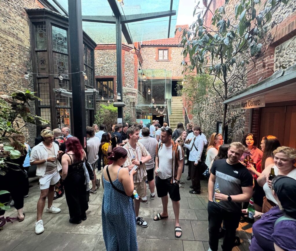 A group of people socializing and standing in a covered, rustic courtyard with brick and stone walls, glass ceiling, plants, and a staircase leading to an upper level. Some hold drinks and appear to be enjoying a casual event.