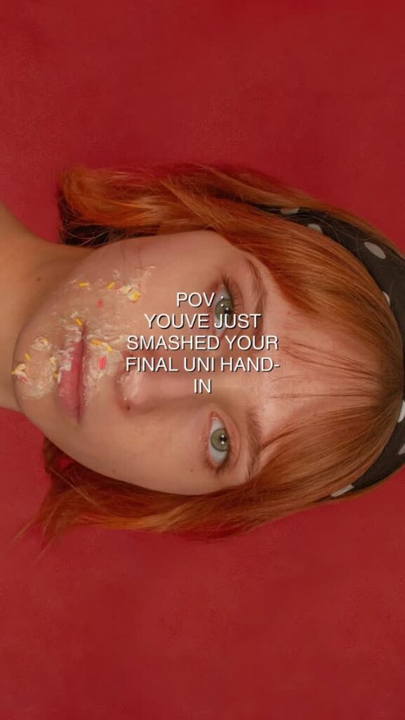 A young person with red hair and a black headband lies on a red surface, cake smeared on their cheek. Text over the image reads: "POV you’ve just smashed your final uni hand-in.