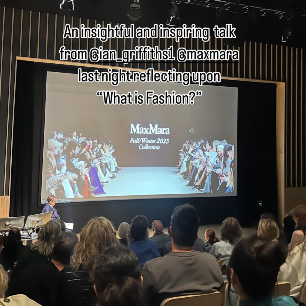 A speaker addresses an audience in a modern auditorium. A large screen displays “MaxMara Fall/Winter 2025 Collection” and images of a runway show, with text above reflecting on the talk: “What is Fashion?”.