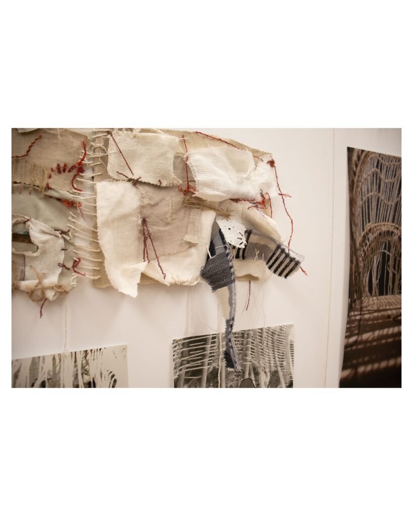 A textile artwork with layered, frayed fabric, red stitching, and patches of gray and striped material is displayed on a white wall above abstract black-and-white prints.