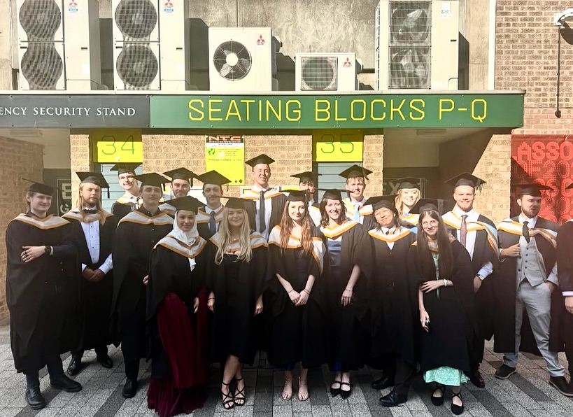 A group of university graduates in caps and gowns stand together outside under signs reading “Seating Blocks P-Q,” smiling and posing for a photo.