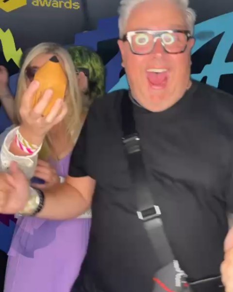 A man in black sunglasses and a black shirt smiles excitedly at the camera, while a woman beside him in a purple dress holds a mango in front of her face. The background features colorful designs and awards text.