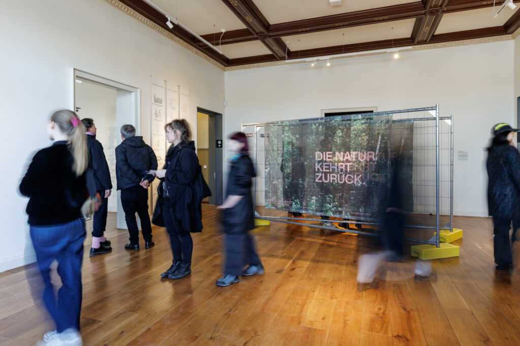 People walk through a modern art gallery with wooden floors. A large display with the German text "DIE NATUR KEHRT ZURÜCK" stands in the center. Some visitors are blurred, suggesting movement.