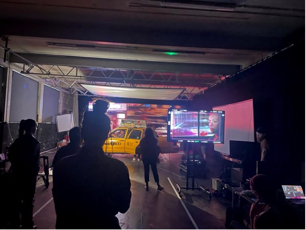 A film set with crew members silhouetted in a dimly lit foreground, a yellow taxi in the background, and monitors displaying a scene with a man’s face and city lights. Equipment and cables are visible around the set.