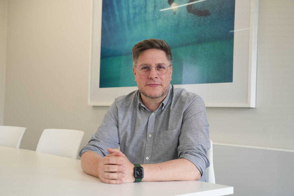 A person with short hair and glasses sits at a white table, wearing a gray shirt and a smartwatch, with a large abstract blue artwork on the wall behind them.